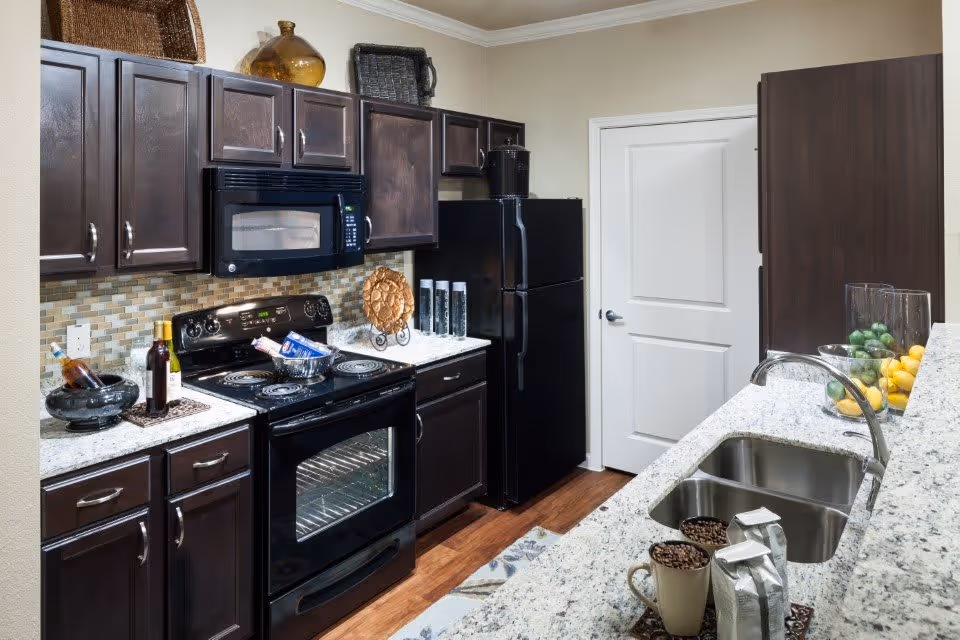 Modern kitchen with dark wood cabinets, black appliances, a granite countertop island with a double sink, and bowls of fruit and coffee items.