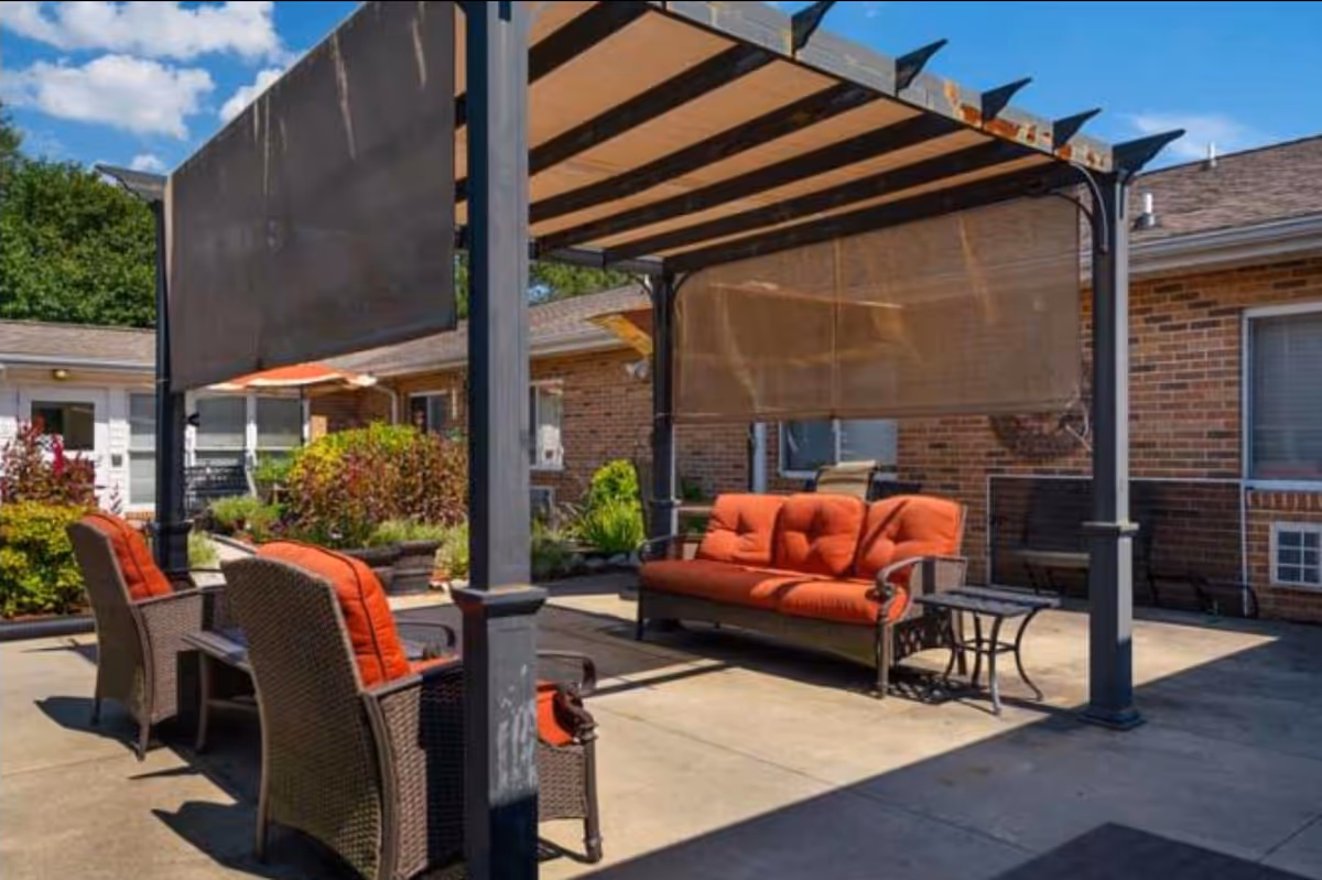 Outdoor patio seating with red cushions under a shaded pergola beside a brick building.