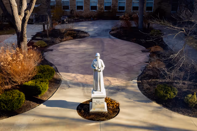 A statue of a robed figure standing on a pedestal in the center of a paved courtyard surrounded by landscaped bushes and trees, with a brick building in the background.