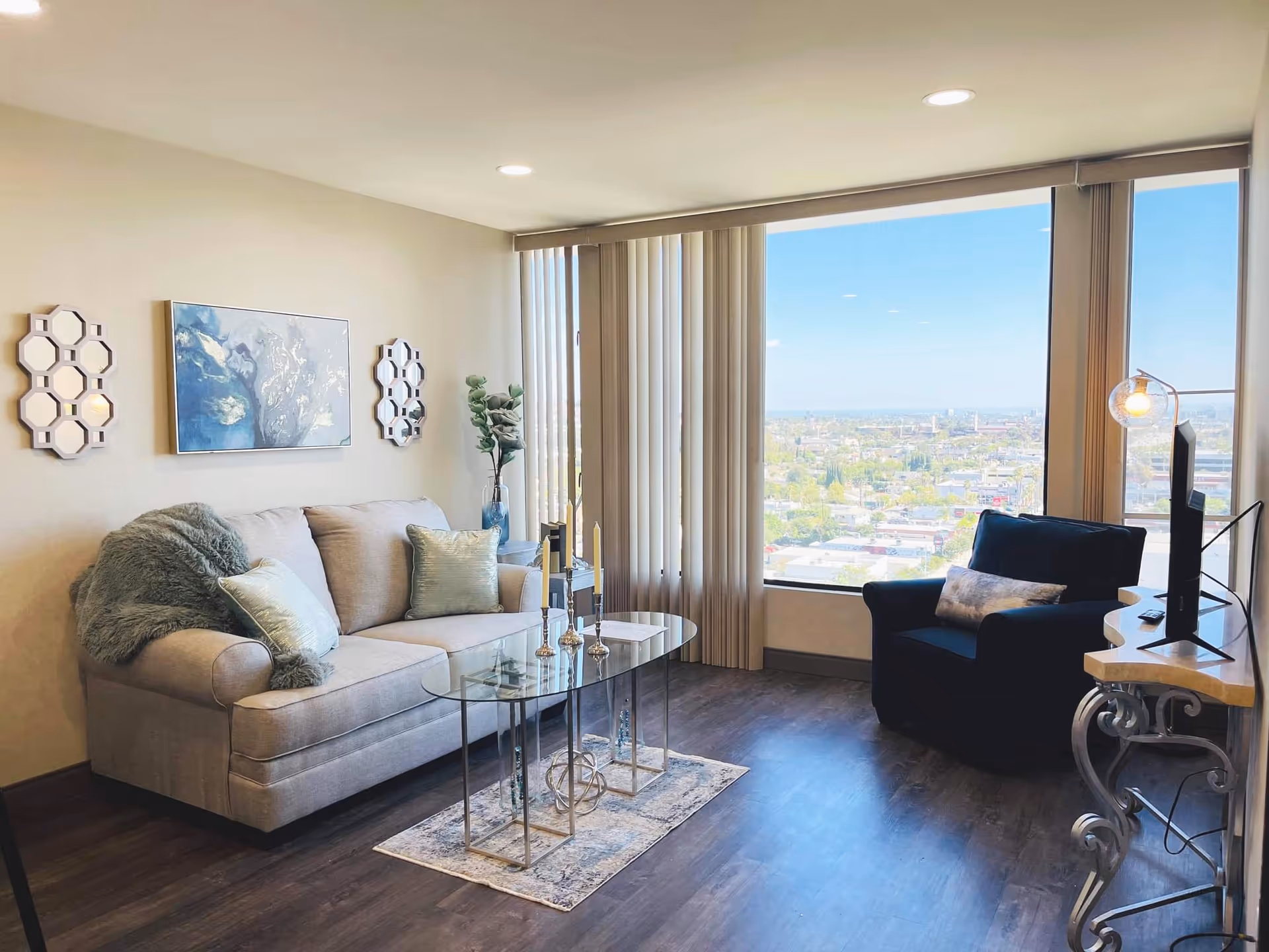A bright living room with a large window offering a city view. The room features a beige sofa with decorative pillows and a throw, a glass coffee table with candles, a dark armchair with a pillow, a TV on a decorative stand, and wall art including two hexagonal mirrors and a painting.