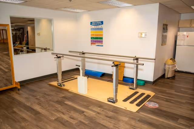 Physical therapy area with parallel gait bars, mirror, exercise steps and a refrigerator in a senior facility.