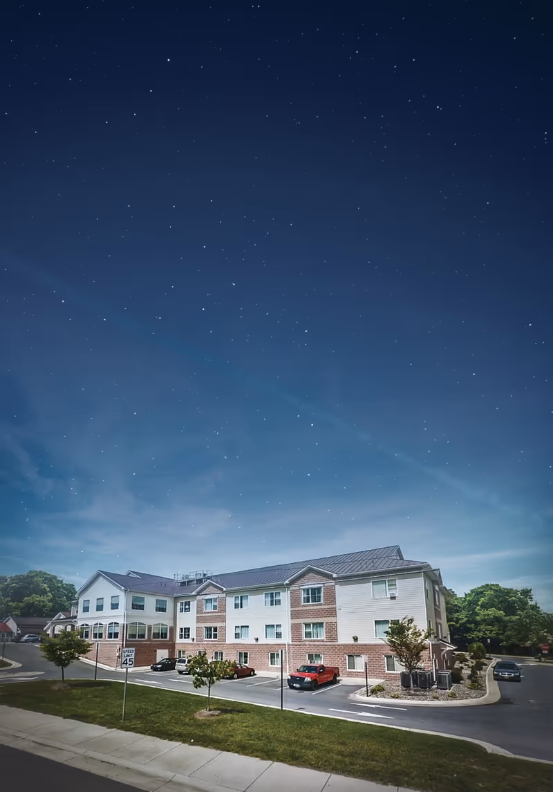 A three-story senior living facility building under a clear night sky filled with stars. The building has a combination of brick and light-colored siding exterior with multiple windows. There is a parking lot in front with several parked cars and small trees along the sidewalk and parking area.
