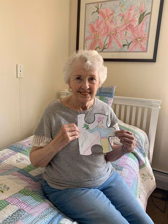 An elderly woman with white hair is sitting on a bed with a colorful patchwork quilt. She is smiling and holding a large puzzle piece with a drawing of a person wearing a hat and holding a plant. Behind her, there is a framed picture of pink lilies on the wall.