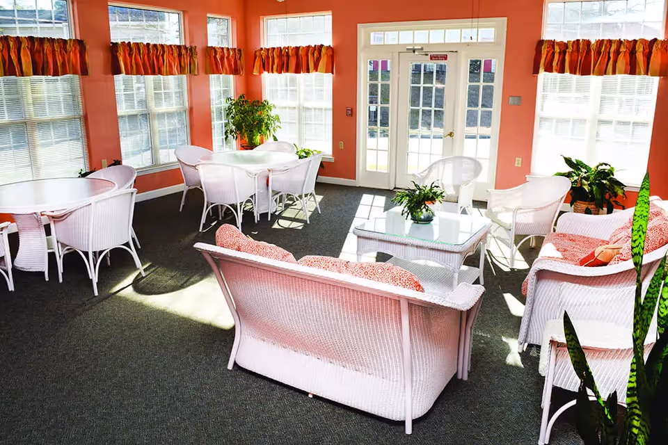 A bright and cozy sitting area with white wicker furniture including chairs, sofas with red patterned cushions, and tables. The room has large windows with blinds and colorful valances, coral-colored walls, and several green plants placed around. Sunlight streams through the windows and glass doors leading outside.