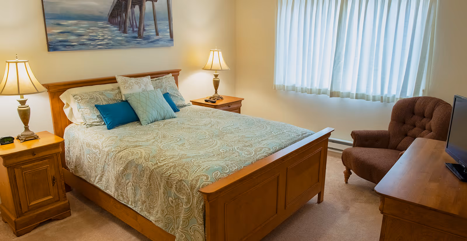 A cozy bedroom with a wooden bed frame, a bed made with patterned bedding and multiple pillows. There are two wooden nightstands with lamps on either side of the bed. A brown upholstered armchair is placed near a wooden dresser with a flat-screen TV on top. A large window with white curtains allows natural light into the room. A painting of a pier over water hangs on the wall above the bed.