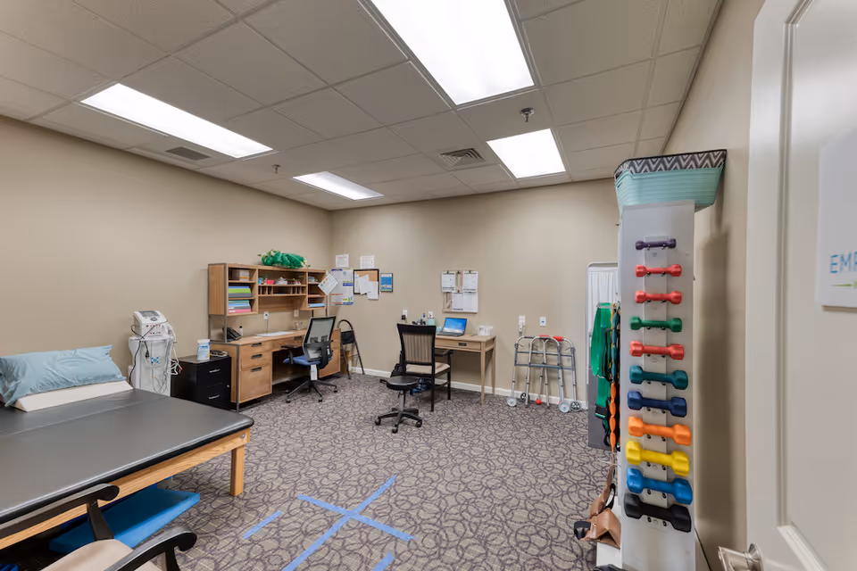 A therapy or rehabilitation room with a padded treatment table on the left, two office desks with chairs against the far wall, a walker, and a rack holding colorful hand weights on the right. The room has beige walls, patterned carpet, and fluorescent ceiling lights.