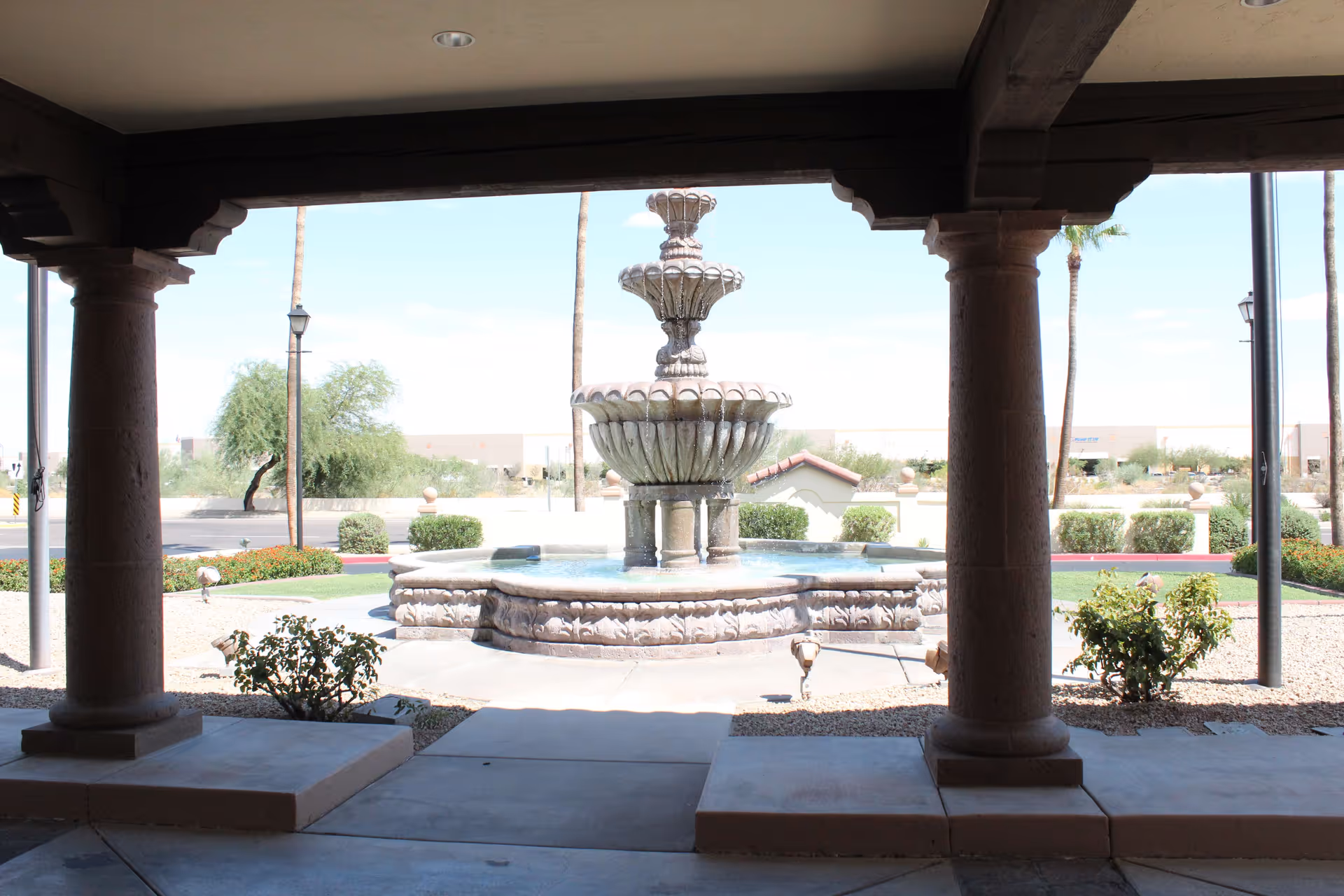 View of a multi-tiered stone fountain outside, framed by large stone columns and a covered patio area. There are bushes and desert landscaping around the fountain, with a clear sky and some palm trees in the background.