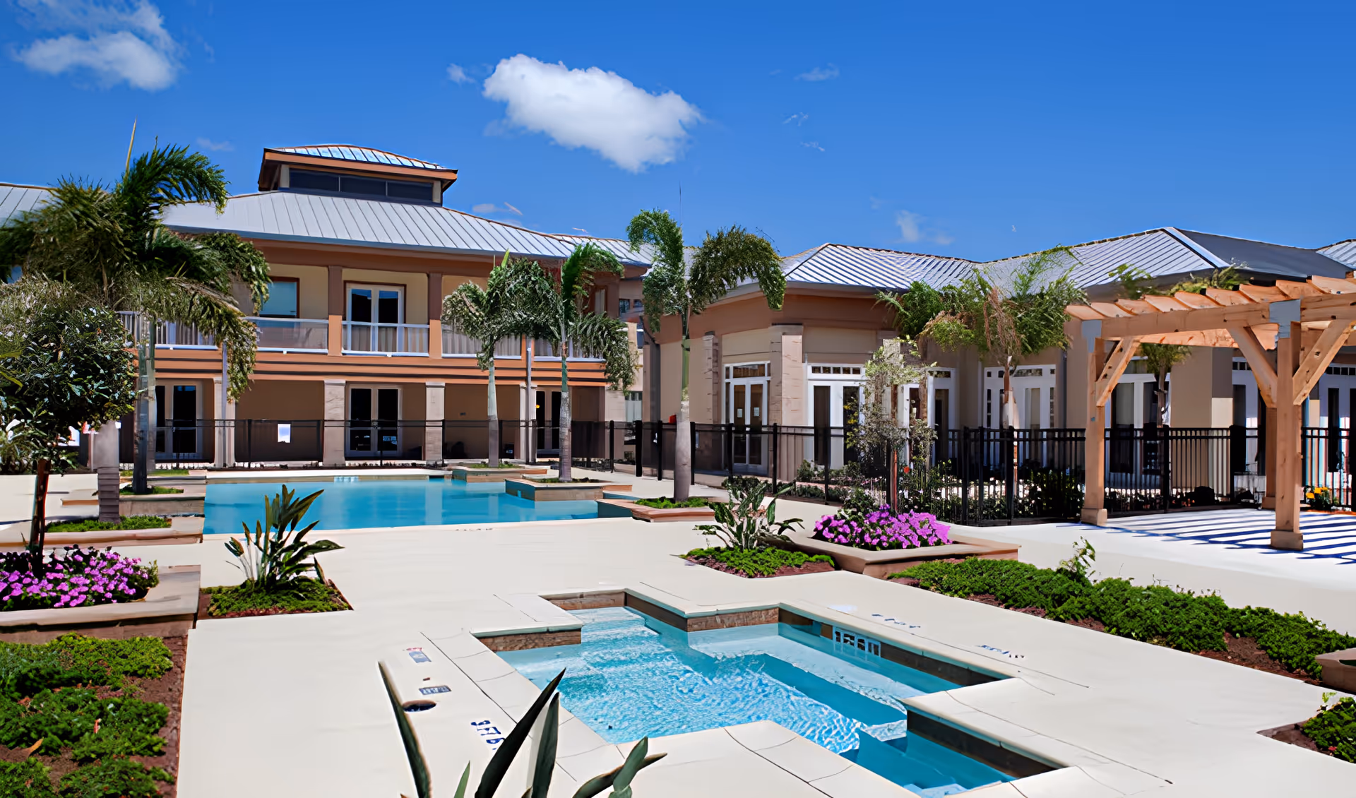 Outdoor courtyard with a swimming pool, hot tub, palm trees, pergola, and a two-story building under a clear blue sky.
