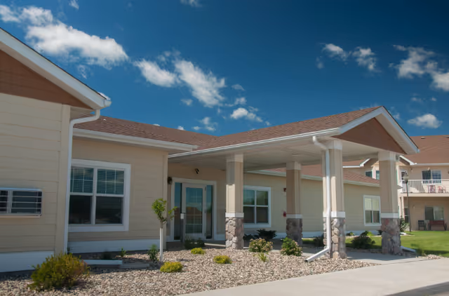 Exterior view of a senior living facility building with beige siding, a covered entrance supported by stone pillars, and a clear blue sky with some clouds.