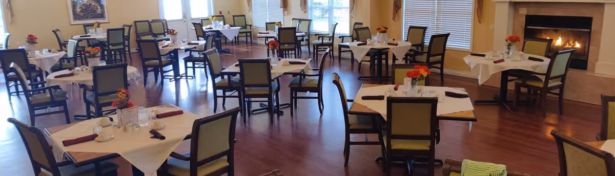 A spacious dining room with multiple square tables covered with white tablecloths and set with cups, glasses, and napkins. Each table has a small floral centerpiece. The room has wooden flooring, large windows with blinds, and a lit fireplace on the right side.