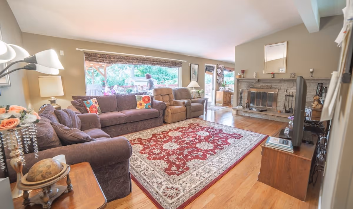 A bright living room with a large red and white patterned area rug on a wooden floor. The room features two purple sofas with colorful floral pillows, a beige recliner chair, a stone fireplace with a mirror above it, and a TV on a wooden stand. There are large windows and a glass door letting in natural light and showing an outdoor patio with people outside.