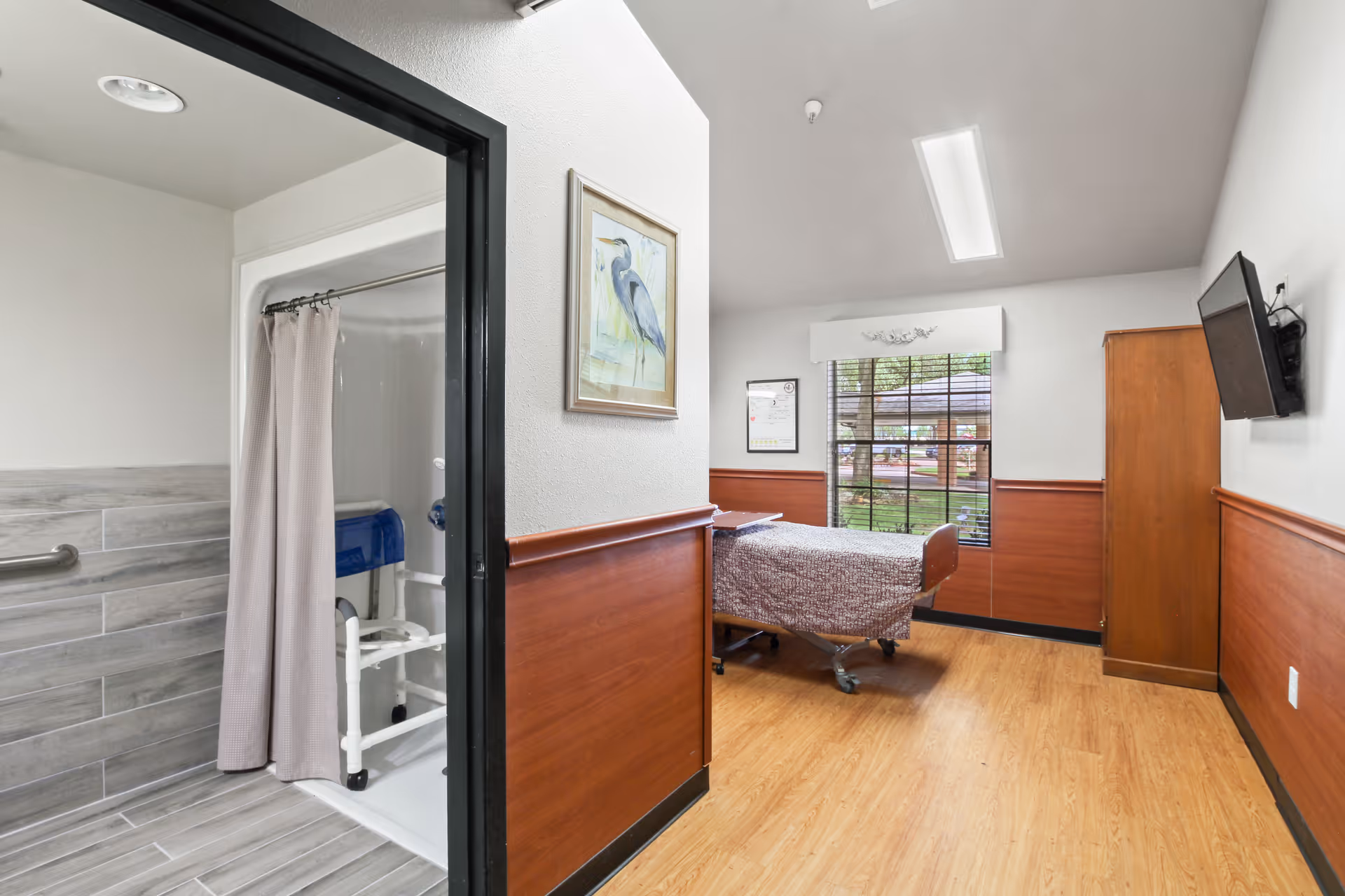 A room in a skilled nursing facility featuring a hospital bed with a patterned blanket, a wooden wardrobe, a wall-mounted TV, and a window with blinds showing an outdoor view. Adjacent to the room is a bathroom with a shower equipped with a shower chair and a curtain. The room has wood paneling on the lower half of the walls and light-colored flooring.