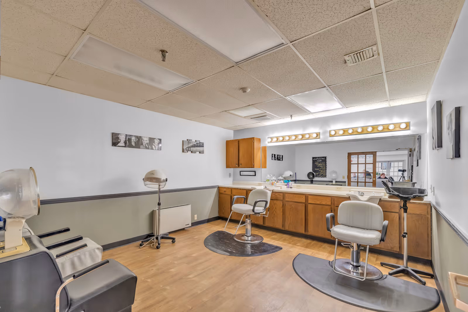 Interior view of a hair salon area in a nursing home with two salon chairs on black mats, a large mirror with lights above it, wooden cabinets below the mirror, hair drying machines, and black and white photos on the walls.