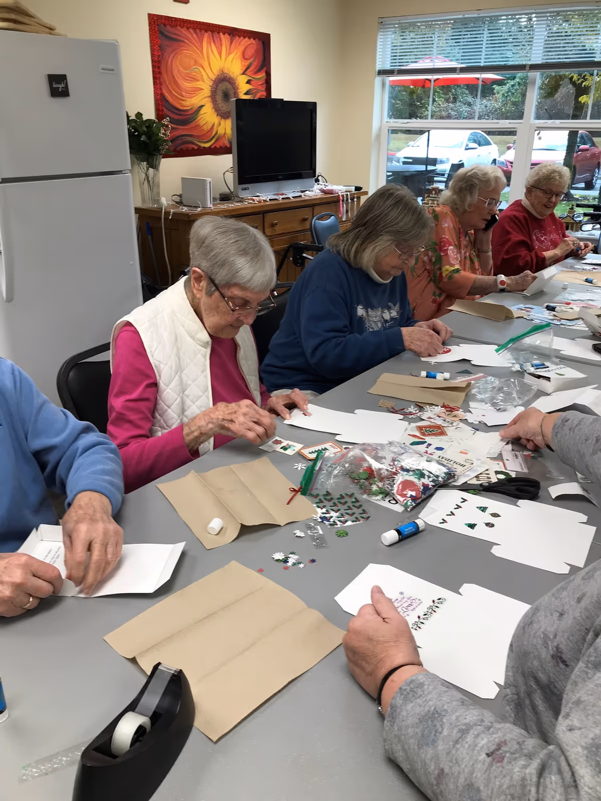 A group of elderly women sitting around a table engaged in arts and crafts activities, decorating paper with stickers and glue. The room has a refrigerator, a TV on a wooden cabinet, a sunflower wall hanging, and a window showing cars and greenery outside.