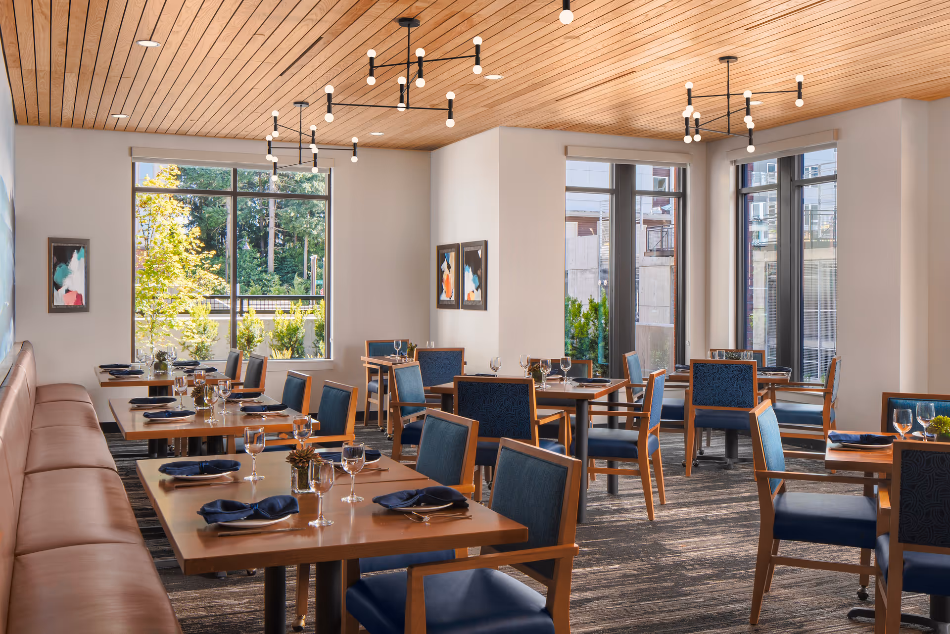 A bright and modern dining room with wooden tables set with wine glasses, plates, and navy blue napkins. The room features blue cushioned chairs with wooden frames, large windows letting in natural light, abstract artwork on the walls, and a wooden ceiling with contemporary light fixtures.