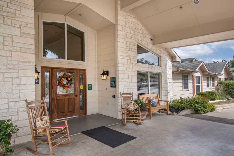 Entrance area of Heritage Place of Boerne featuring a wooden door decorated with a fall-themed wreath, two wooden rocking chairs with cushions, a wooden bench with pillows, stone walls, and a covered porch with outdoor lighting.