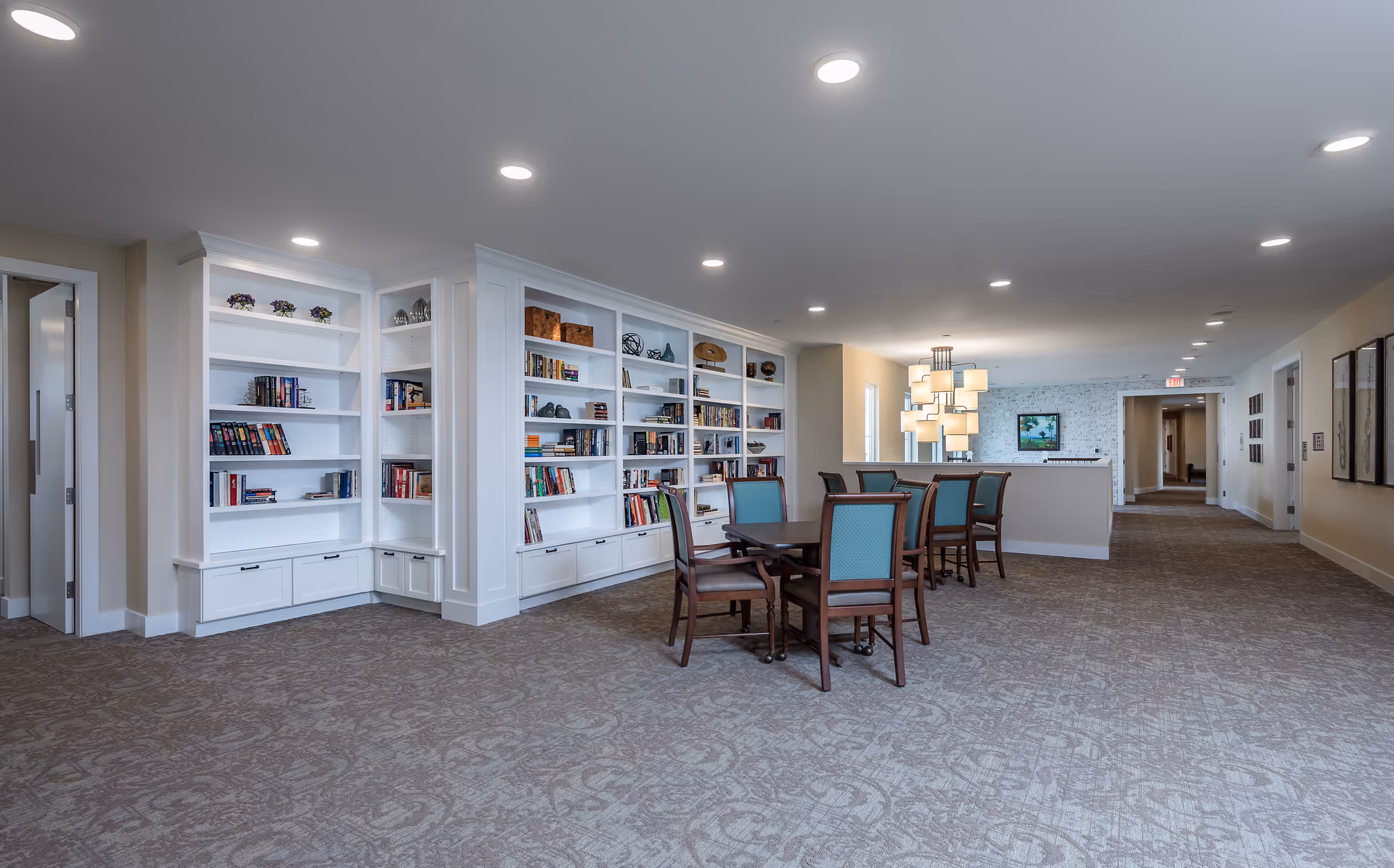 A spacious and well-lit common area with built-in white bookshelves filled with books and decorative items. There are two tables with wooden chairs that have teal upholstery. The room has beige walls, patterned carpet flooring, recessed ceiling lights, and a modern chandelier hanging above one of the tables. A hallway extends from the room with framed artwork on the walls.