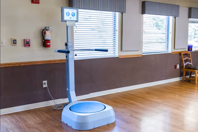 A vibration-platform exercise machine sits on a wood floor in a bright community room with windows and a chair.