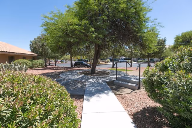 Outdoor area at La Cañada Care Center featuring a concrete pathway leading to a circular seating area under a large tree, surrounded by bushes and desert landscaping with gravel. A parking lot with cars is visible in the background under a clear blue sky.