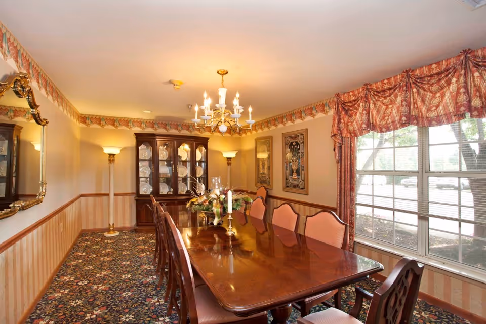 A traditional dining room with a long polished wooden table surrounded by upholstered chairs. The room features a patterned carpet, a chandelier hanging from the ceiling, two floor lamps, a large window with red patterned curtains, a wooden china cabinet displaying plates, and framed artwork on the walls.