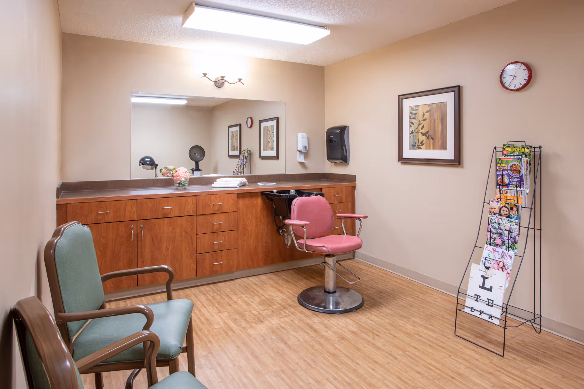 A small salon or grooming room with a pink salon chair in front of a counter with wooden cabinets and a large mirror. Two green chairs are placed against the opposite wall. A magazine rack filled with magazines stands near a framed picture and a clock on the beige wall. The floor is wood-patterned.