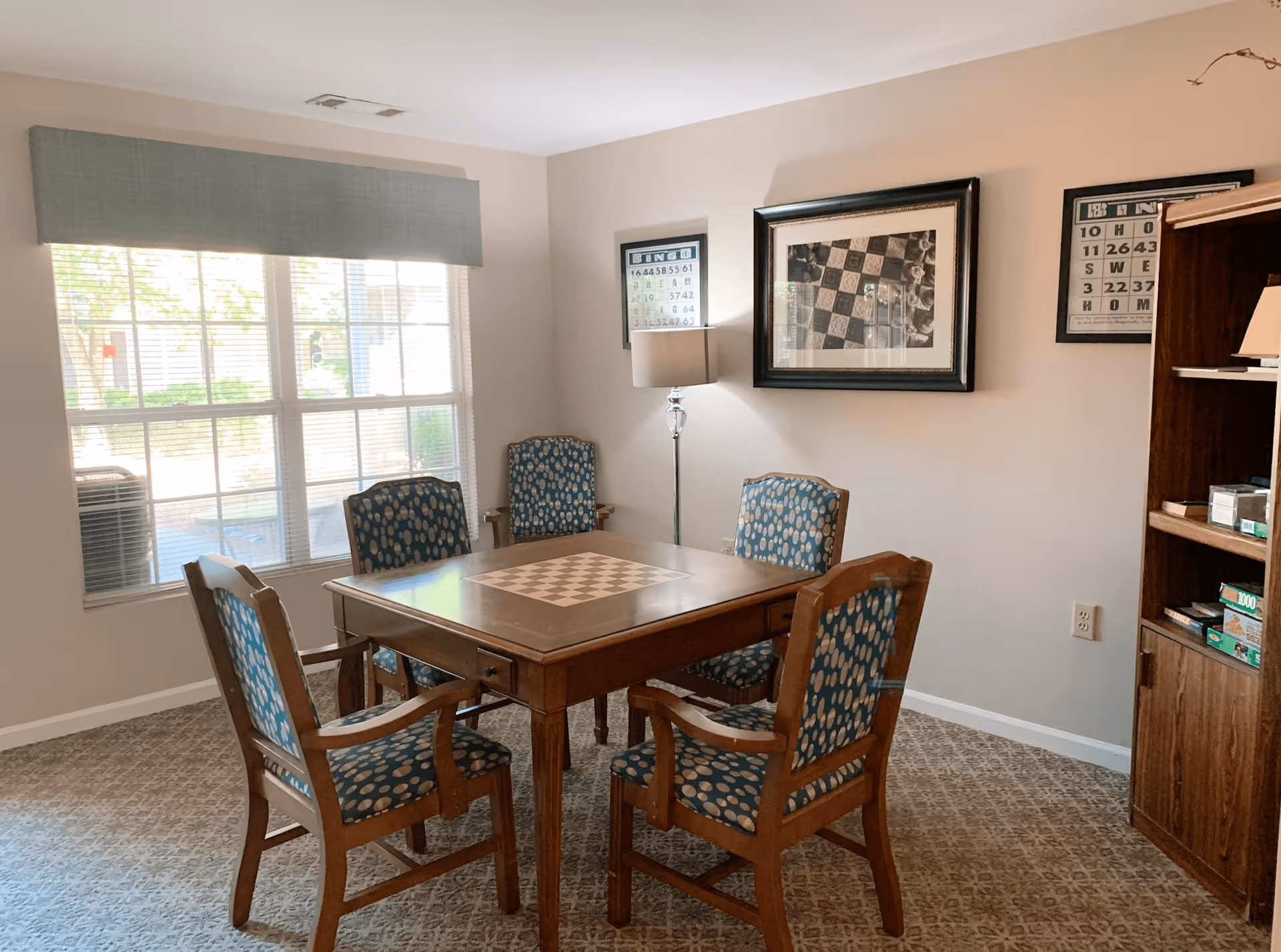 A well-lit room with a wooden table featuring a built-in chessboard, surrounded by five wooden chairs with blue patterned cushions. The room has a large window with blinds, a floor lamp, framed artwork on the walls, and a wooden cabinet with board games and books.