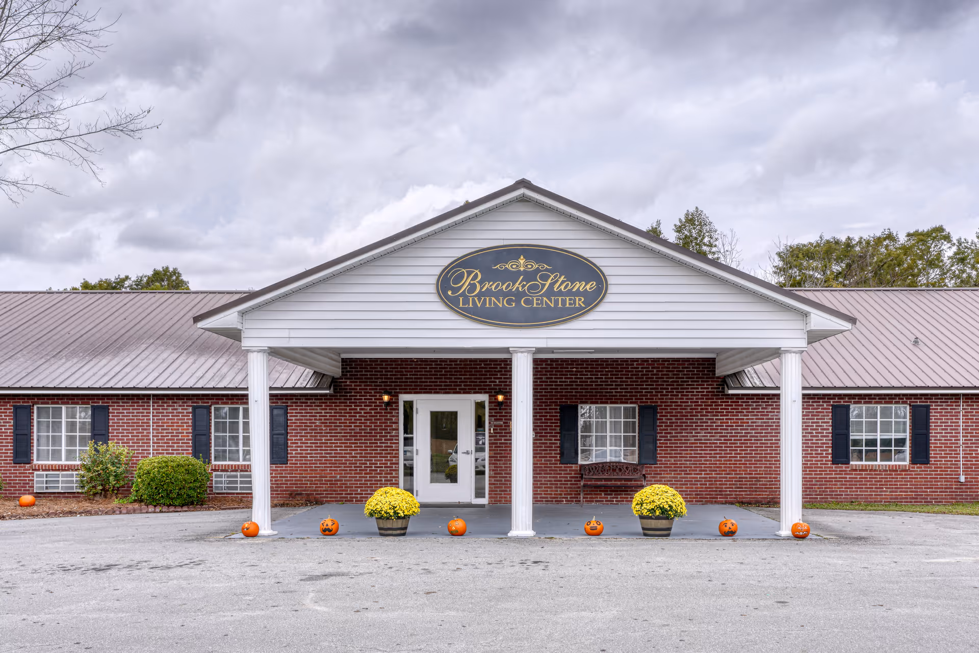 Front exterior view of Brook Stone Living Center, a single-story brick building with white columns supporting a covered entrance. The sign above the entrance reads 'Brook Stone Living Center'. There are pumpkins and yellow potted flowers decorating the entrance area, and the sky is cloudy.