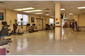 A spacious interior room in a senior living facility with exercise equipment such as stationary bikes and treadmills along the walls. The room has a tiled floor, fluorescent ceiling lights, and windows letting in natural light. There are chairs and tables near the windows and a hallway leading to other areas of the facility.
