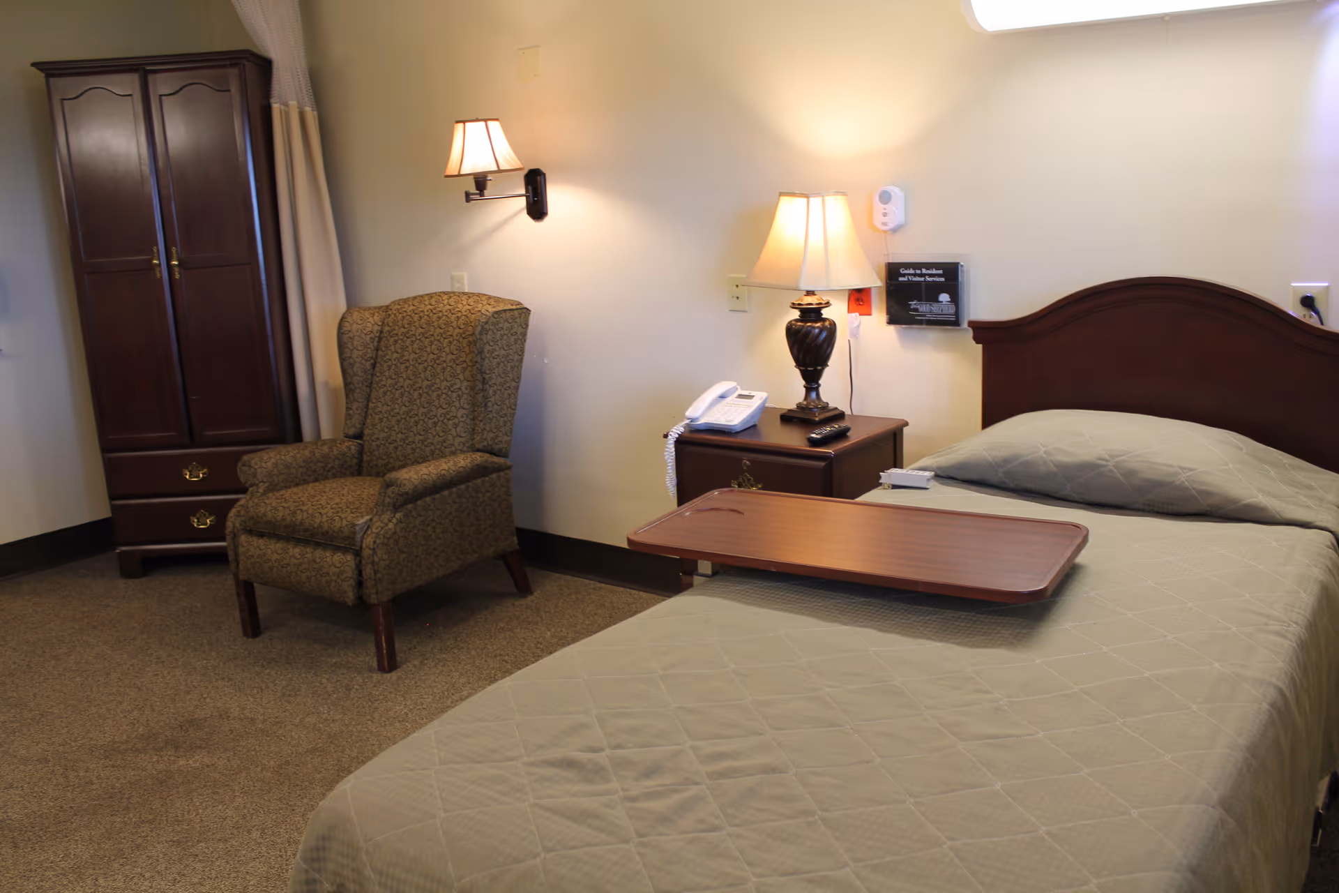 A neatly arranged nursing facility bedroom with a single bed and tray, nightstand with lamp and phone, upholstered armchair, and wooden wardrobe.