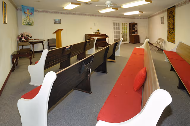 Interior room with wooden pews featuring red cushions arranged in rows facing a small podium and a piano. The room has white walls decorated with religious artwork and a floral arrangement on a side table. There is a ceiling fan and fluorescent lighting overhead.