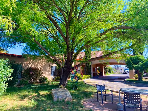 Outdoor view of Desert Peaks Assisted Living and Memory Care facility showing a large tree with green leaves, a stone bench, patio table with chairs, and part of the building with a covered entrance and driveway.