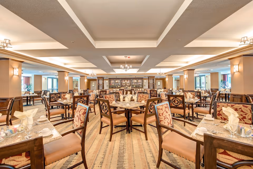 A spacious and well-lit dining room with multiple tables and chairs arranged neatly. Each table is set with napkins, cups, and utensils, and the chairs have patterned upholstery. The room features a coffered ceiling with recessed lighting and chandeliers, beige walls with wooden accents, and large windows allowing natural light to enter.