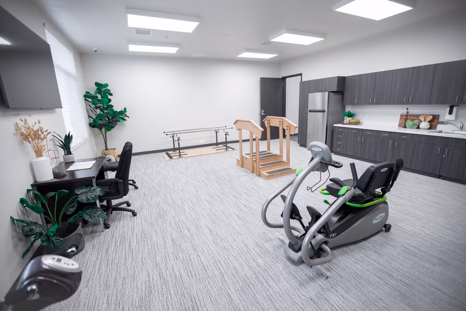 A spacious rehabilitation room with exercise equipment including a recumbent bike and wooden parallel bars for physical therapy. The room has gray carpet, white walls, a desk with a chair, several potted plants, and a kitchenette area with dark cabinets and a stainless steel refrigerator. Bright ceiling lights illuminate the clean and organized space.