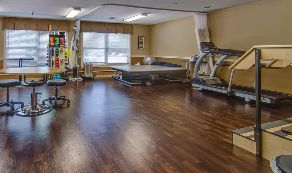 A spacious room with wooden flooring featuring exercise equipment including a treadmill, therapy table, exercise balls, and a rack with various colorful exercise bands and weights. There are two windows with blinds and a round table with stools on the left side.