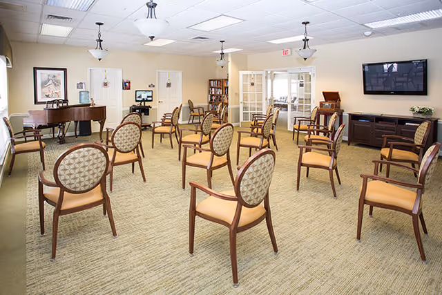 Bright communal activity room with evenly spaced upholstered chairs, a piano, a television, and bookshelves.