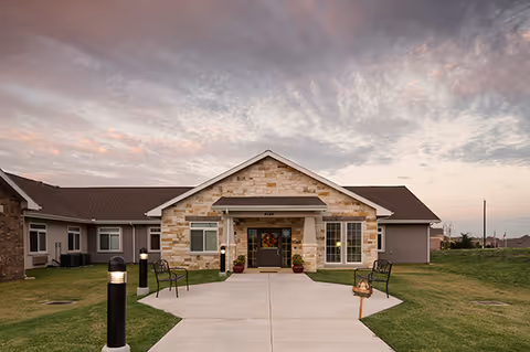 Front exterior view of a single-story building with stone and siding facade, a walkway leading to the entrance, outdoor benches, and lamp posts on a grassy lawn under a cloudy sky at sunset.