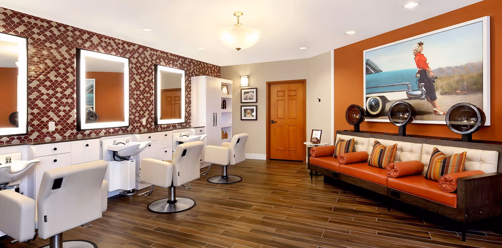 Interior view of a salon area in a senior living facility with three white salon chairs in front of mirrors and sinks. The wall behind the chairs has a decorative red and white patterned wallpaper. To the right, there is a seating area with a long orange and white cushioned bench, striped pillows, and three vintage hair dryers mounted on the wall. A large painting of a woman standing next to a vintage car hangs above the bench. The floor is wooden, and there is a wooden door in the background.