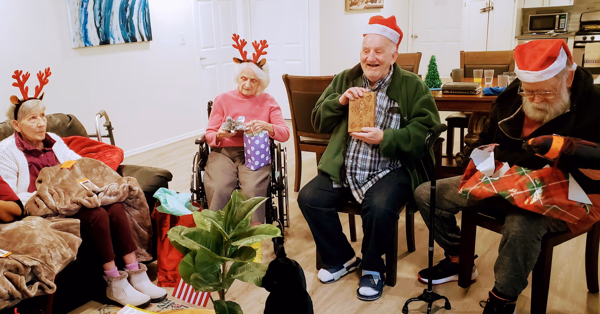 Four elderly people sitting in a living room decorated for Christmas, wearing festive accessories like reindeer antlers and Santa hats, opening gifts and smiling. The room has a dining table and kitchen appliances in the background.