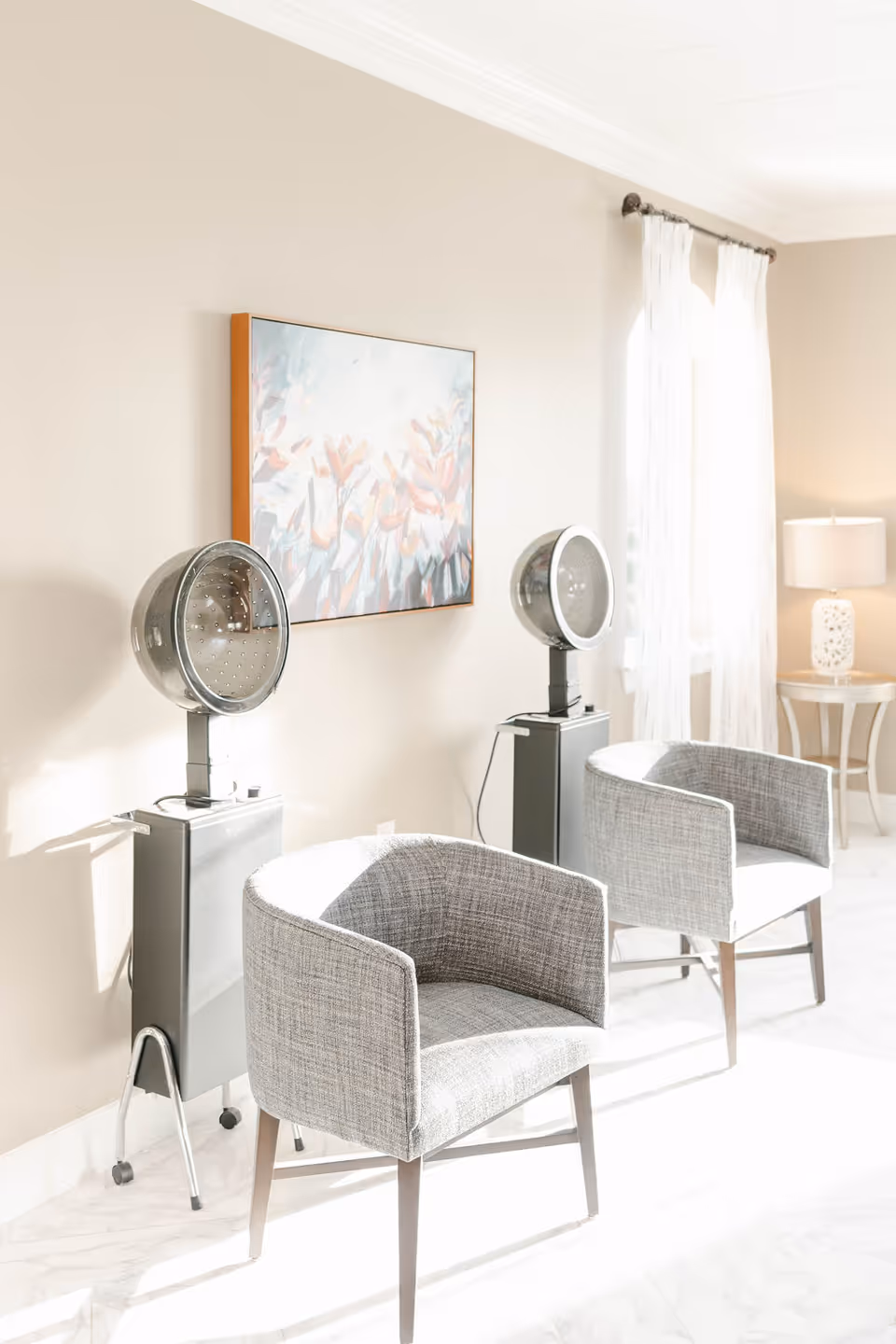 A bright room with two gray upholstered chairs positioned in front of two vintage hair dryer stations. A colorful floral painting hangs on the beige wall above the hair dryers. Sheer white curtains cover a window, and a small round table with a white lamp is visible in the background.