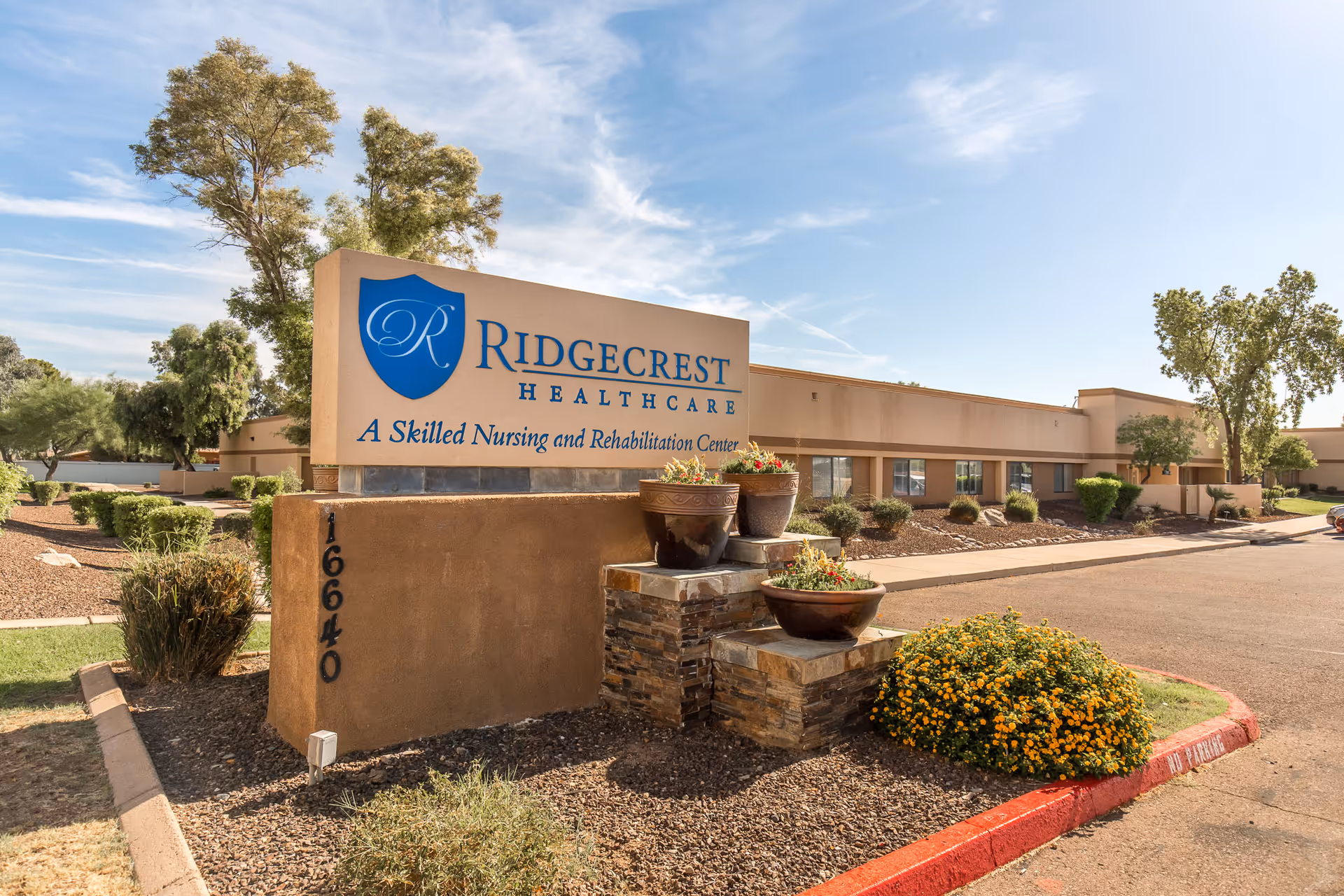 Exterior view of Ridgecrest Healthcare, a skilled nursing and rehabilitation center, showing the building and a large sign with the facility's name and address surrounded by landscaping and trees under a partly cloudy sky.