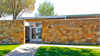 Exterior view of Sunset Villa Care Center showing a stone wall facade with the facility name displayed. There is a glass entrance door in the center with trees and green grass surrounding the pathway leading to the entrance.