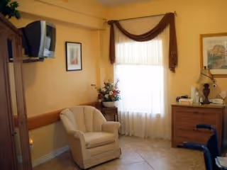 A cozy room with a beige armchair, a wooden dresser with a lamp and decorative items, a window with sheer white curtains and a brown valance, a wall-mounted TV, and framed artwork on the walls.