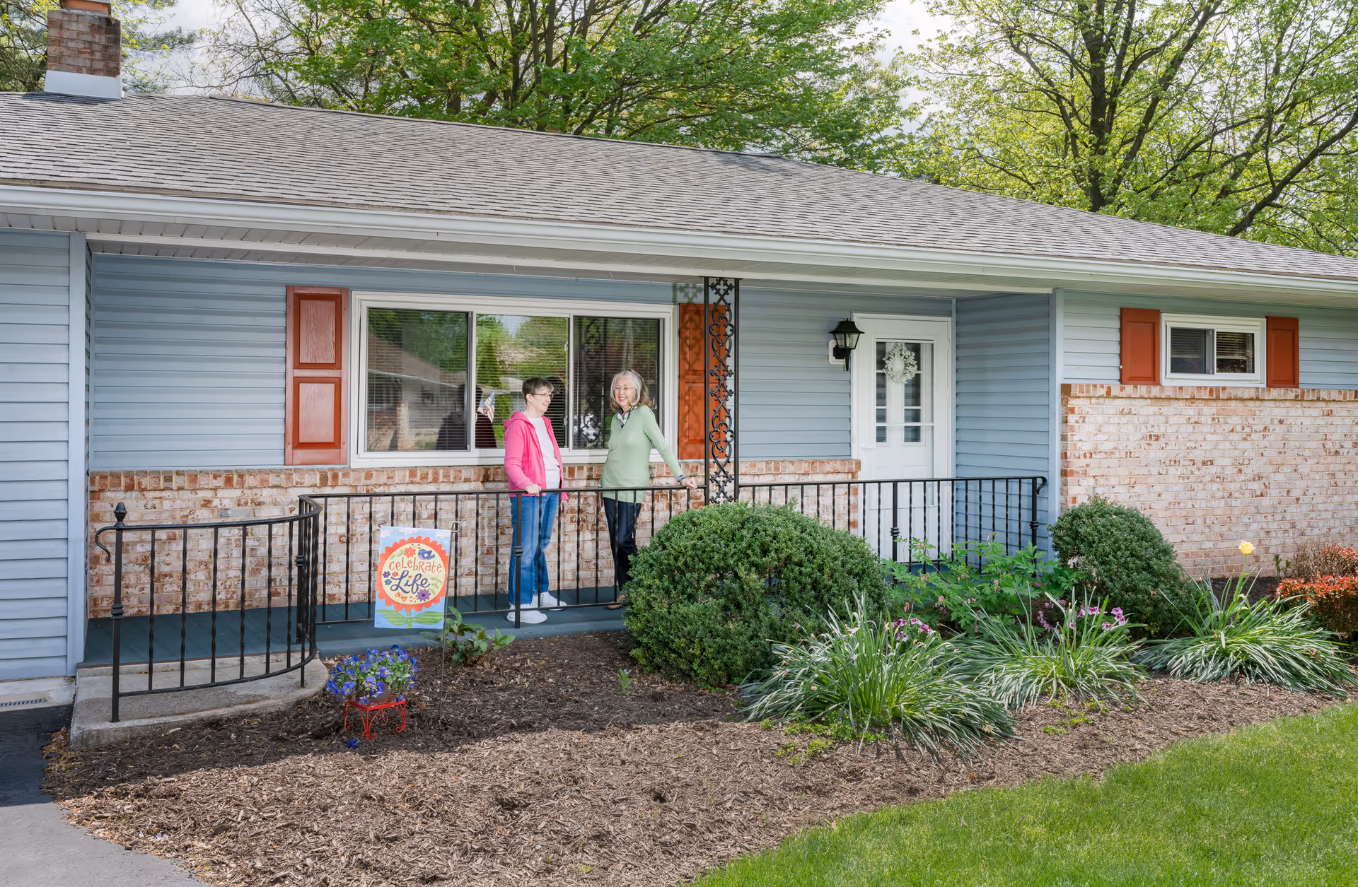 Two women stand on the front porch of a single-story house with blue siding, brick accents, a small garden, and a decorative sign.
