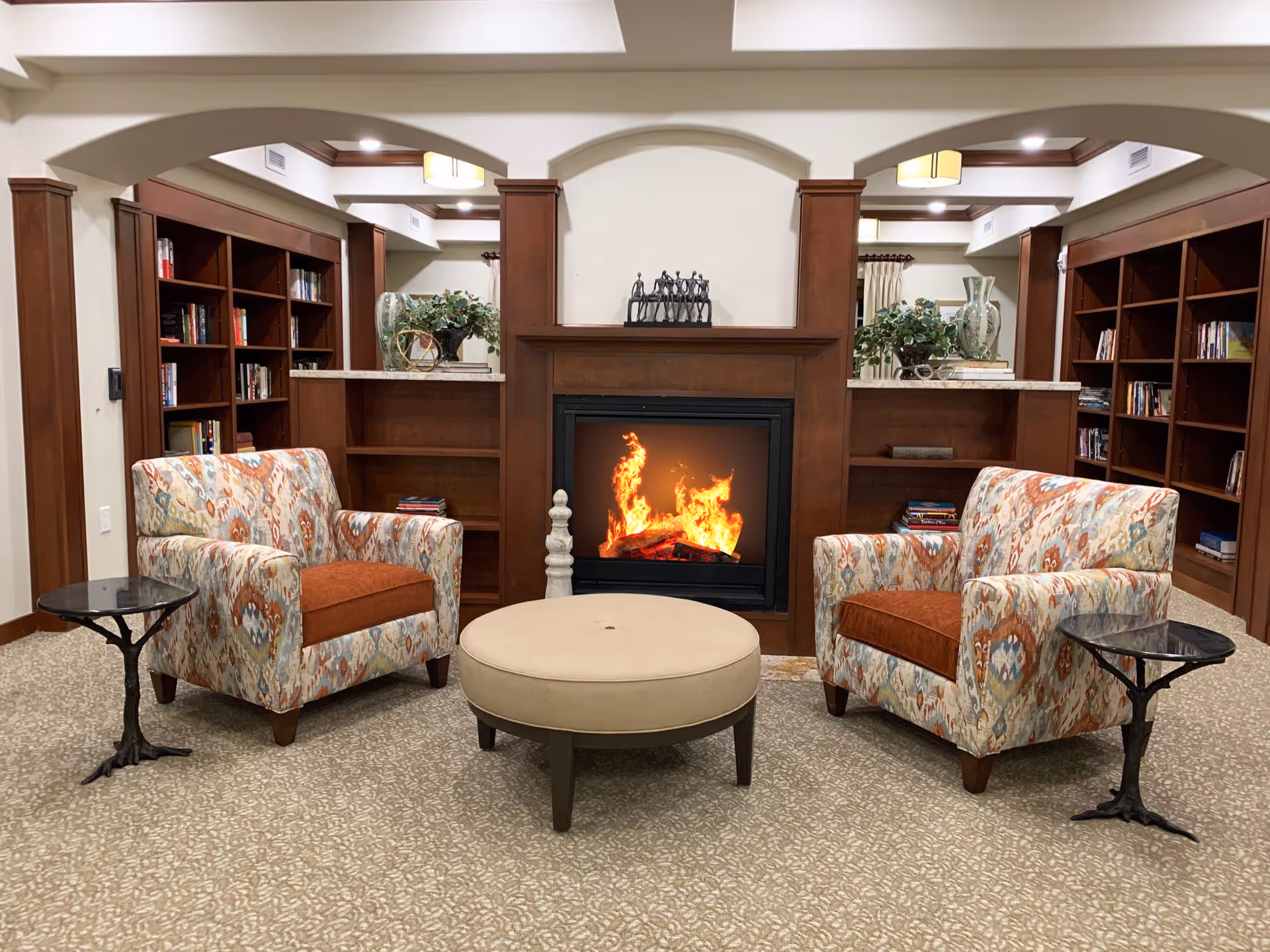 A cozy seating area in a retirement community featuring two patterned armchairs with orange cushions facing a round beige ottoman. Behind the ottoman is a wooden fireplace with a fire burning. On either side of the fireplace are built-in wooden bookshelves filled with books and decorative items. The room has a beige patterned carpet and soft lighting.