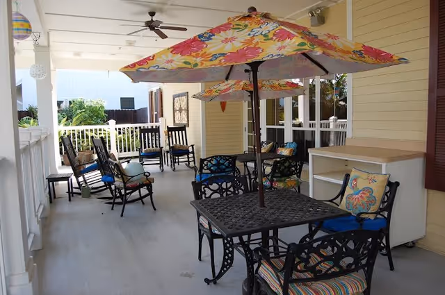 Covered outdoor patio area with multiple black metal chairs and tables. Some tables have colorful floral umbrellas. Cushions with various patterns are on the chairs. Ceiling fans are mounted on the white ceiling. The patio is adjacent to a yellow building with windows and a white railing along the edge.