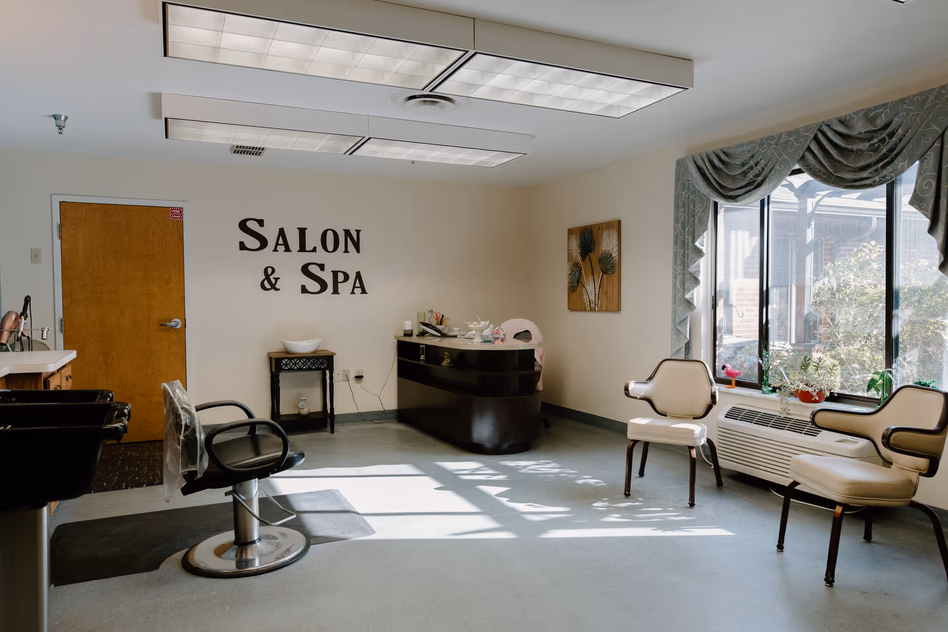 Interior of a salon and spa room with a salon chair, two beige chairs near a large window with green drapes, a small black table with a white bowl, a reception desk, and a wall sign that reads 'SALON & SPA'.