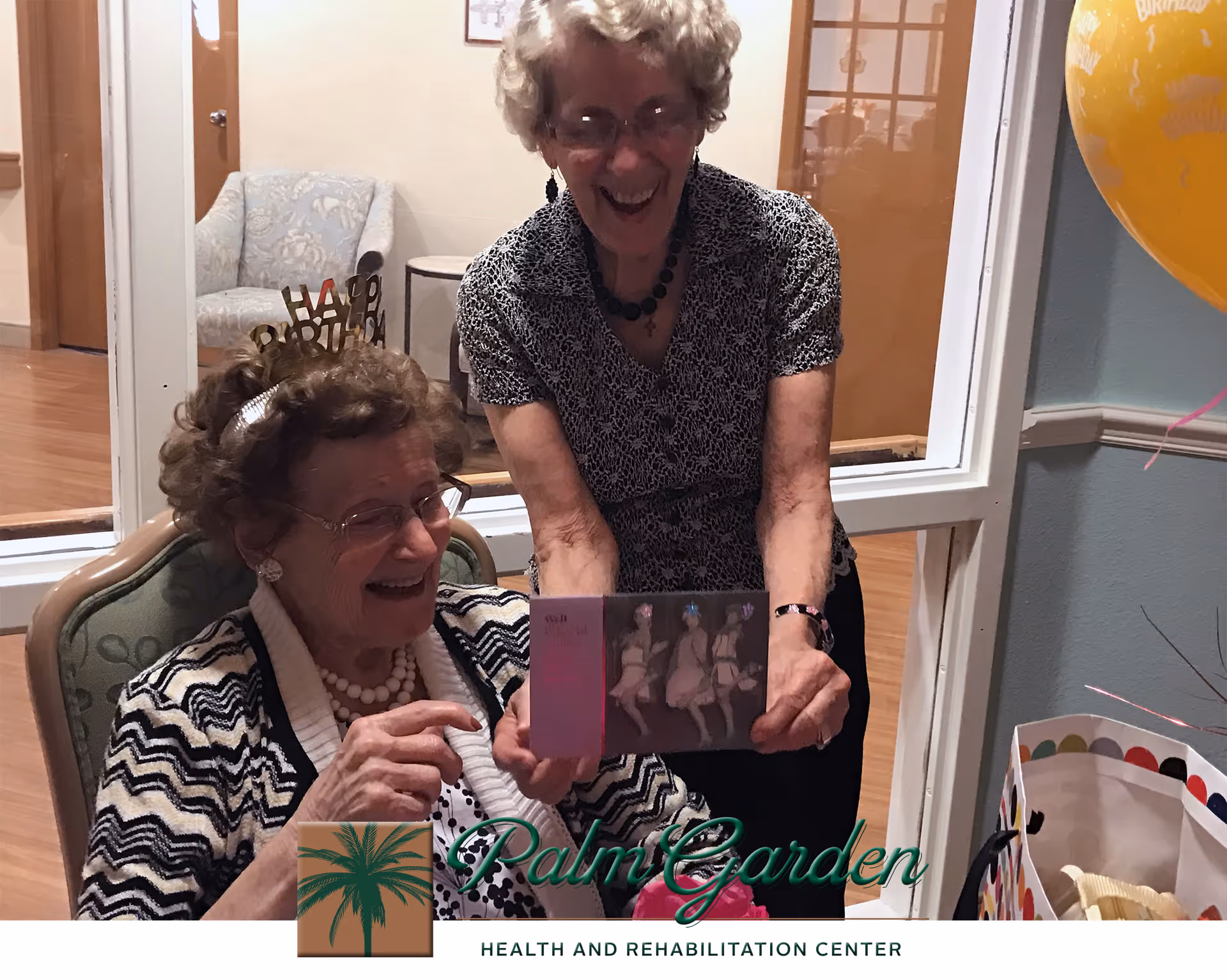 Two elderly women smiling and celebrating a birthday indoors. One woman is seated wearing a 'Happy Birthday' headband and a striped cardigan, while the other woman stands beside her holding a card. There are balloons and a gift bag nearby.