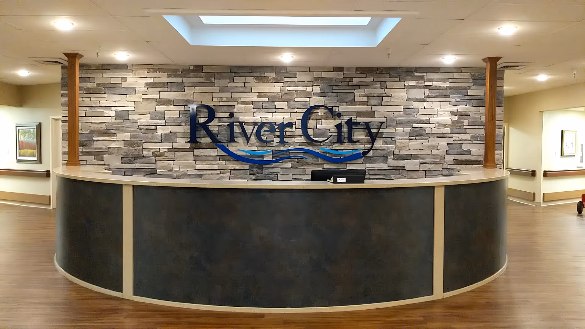 Reception desk area with a curved counter in front of a stone accent wall featuring the sign 'River City' with blue wave designs underneath. The area has wooden flooring and light-colored walls with framed artwork visible in the hallways on either side.