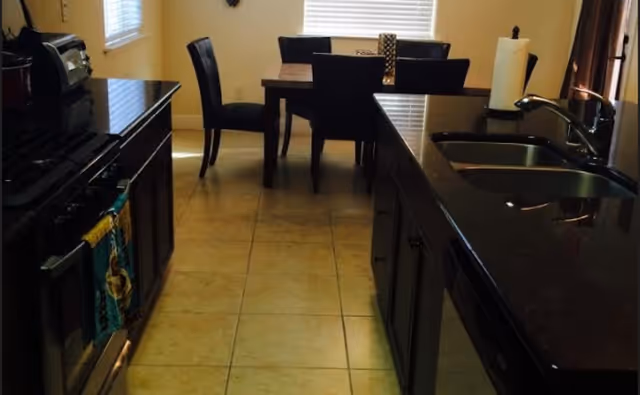 Interior view of a kitchen with dark countertops and a double sink, a stove on the left, tiled floor, and a dining table with chairs by a window.