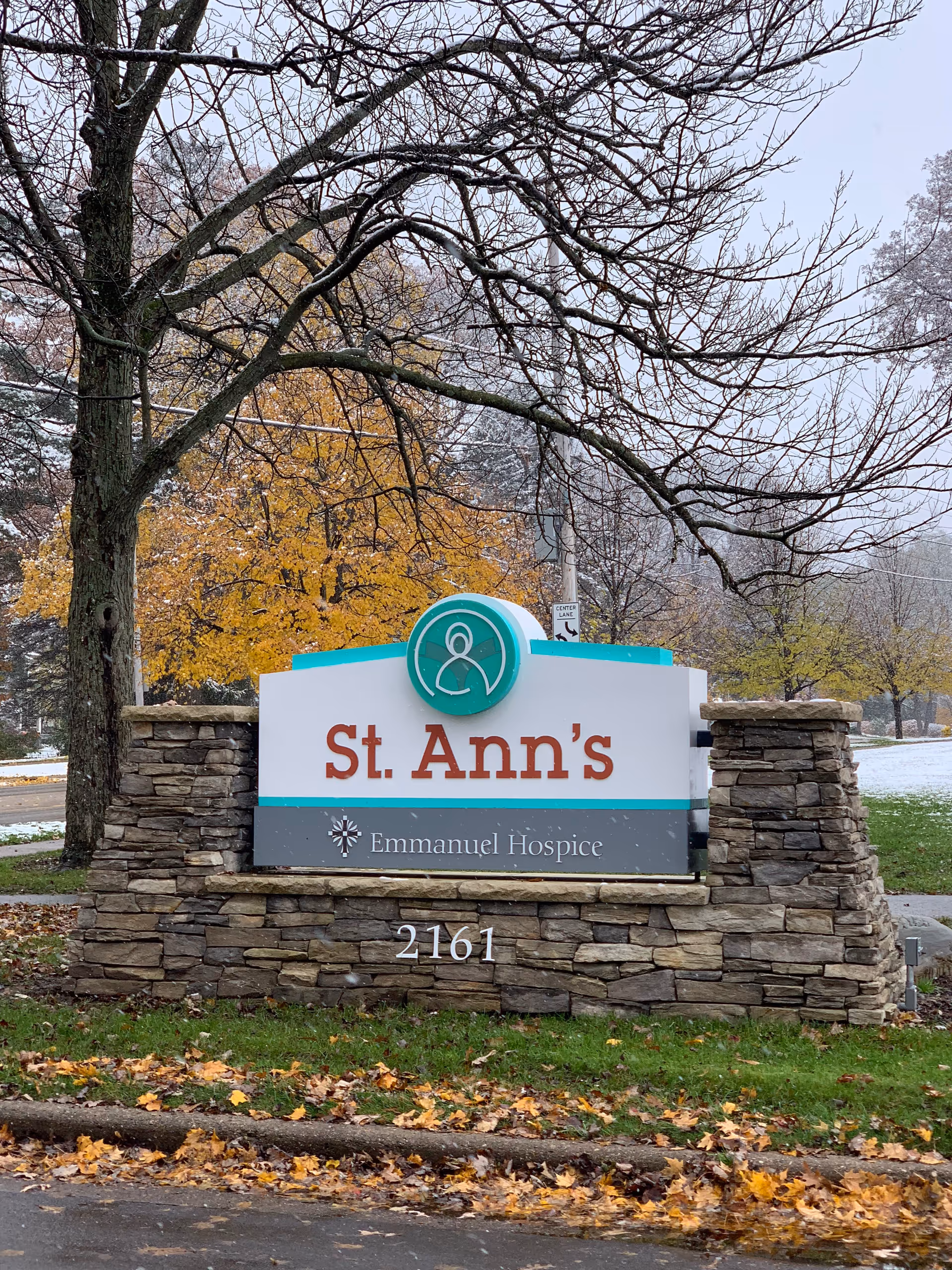 Outdoor stone sign for St. Ann's Emmanuel Hospice with the address number 2161, surrounded by trees with autumn leaves and some snow on the ground.
